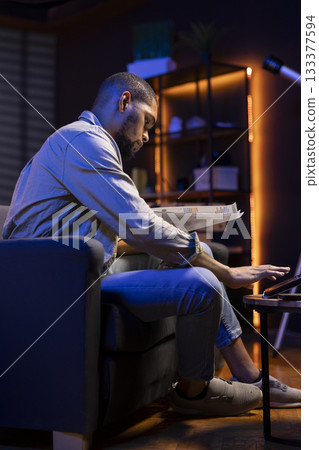 African american freelancer does his research by reading articles and data driven papers in the living room, using online sources to collect information for a new project. Multitasking. 133377594