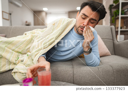 Sick man having a runny nose while lying on a sofa covered with blanket Sick man having a runny nose while lying on a sofa covered with blanket 133377923