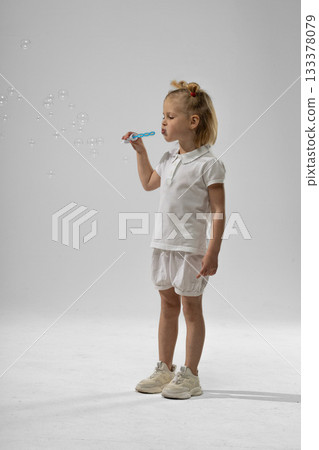 little blonde girl stands happily blowing soap bubbles with a wand. Her playful expression reflects joy as bubbles float around her creating a magical atmosphere. little blonde girl stands happily blowing soap bubbles with a wand. Her playful expression reflects joy as bubbles float around her creating a magical atmosphere. 133378079