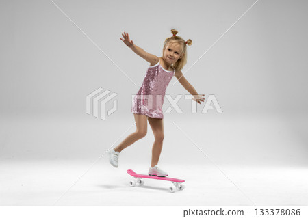 small blonde girl with pigtails balances on a skateboard while wearing a sparkling pink dress. The studio background is simple allowing her joy and energy to stand out. 133378086