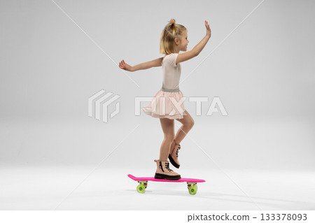 small blonde girl skillfully rides a skateboard wearing a dress and striking a playful pose while balancing on one foot. 133378093