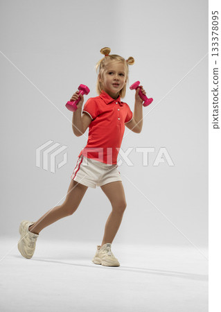 Blonde girl in a red shirt and white shorts smiles as she holds pink dumbbells in each hand. She appears to enjoy exercising in a well lit studio space promoting fitness. Blonde girl in a red shirt and white shorts smiles as she holds pink dumbbells in each hand. She appears to enjoy exercising in a well lit studio space promoting fitness. 133378095