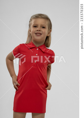 cheerful blonde girl stands in a red dress with a playful smile in a studio. Her expression shows joy as she poses confidently showcasing her outfit against a neutral backdrop. 133378103