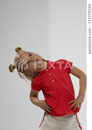 little blonde girl stands with her hands on her hips looking up with a curious expression. She wears a red shirt and has playful hair buns showcasing her joyful spirit. 133378104