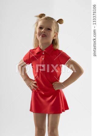 little blonde girl stands confidently with her hands on her hips. She wears a vibrant red dress playfully sticking out her tongue showcasing her personality and energy. little blonde girl stands confidently with her hands on her hips. She wears a vibrant red dress playfully sticking out her tongue showcasing her personality and energy. 133378106