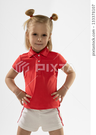 little blonde girl stands confidently with her hands on her hips. She wears a red shirt and white shorts showcasing her playful attitude and joyful expression. 133378107