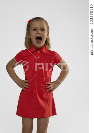 little blonde girl is in a bright studio wearing a red dress. She shows a big smile and appears to be having fun during the session full of energy and joy. 133378110