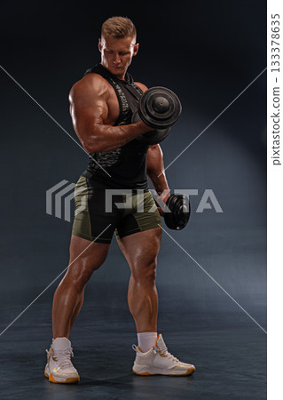 muscular man works out in a gym focusing on strength training with a dumbbell. His posture shows concentration as he lifts weights in a dimly lit space. muscular man works out in a gym focusing on strength training with a dumbbell. His posture shows concentration as he lifts weights in a dimly lit space. 133378635