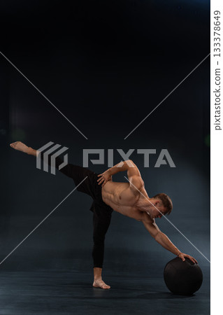 shirtless male athlete balances on one leg while reaching for a large sphere on the floor. The dimly lit training studio creates an atmosphere focused on strength and skill development. shirtless male athlete balances on one leg while reaching for a large sphere on the floor. The dimly lit training studio creates an atmosphere focused on strength and skill development. 133378649