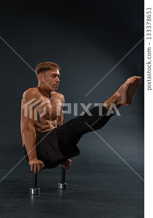 muscular man is executing a challenging leg lift on parallel bars in a well lit training space. His focused expression shows the intensity of his workout. 133378651
