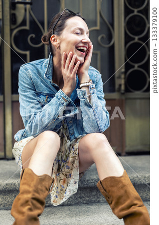 Portrait of young adult woman wear floral dress denim jacket brown suede boots sitting on stairs outdoors in old European city. Stylish female person enjoy walk travel urban lifestyle summertime day Portrait of young adult woman wear floral dress denim jacket brown suede boots sitting on stairs outdoors in old European city. Stylish female person enjoy walk travel urban lifestyle summertime day 133379100