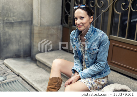Portrait of young adult woman wear floral dress denim jacket brown suede boots sitting on stairs outdoors in old European city. Stylish female person enjoy walk travel urban lifestyle summertime day Portrait of young adult woman wear floral dress denim jacket brown suede boots sitting on stairs outdoors in old European city. Stylish female person enjoy walk travel urban lifestyle summertime day 133379101