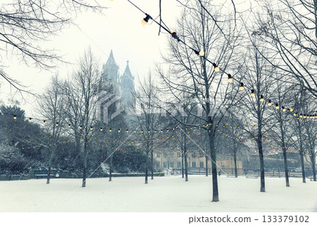 Snow falls over Magdeburg Cathedral, partly hidden behind bare winter trees and glowing garlands of yellow lights. The soft fog and quiet ambiance create a dreamy festive city atmosphere 133379102