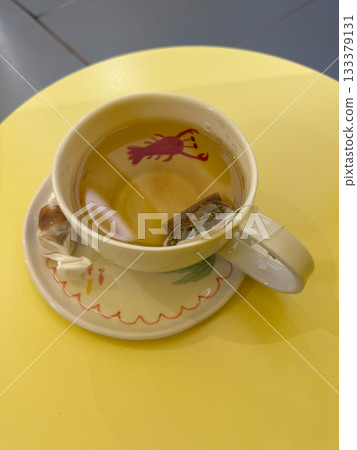 A cup of herbal tea with a tea bag sits on a yellow table, decorated with a whimsical red lobster illustration inside the cup A cup of herbal tea with a tea bag sits on a yellow table, decorated with a whimsical red lobster illustration inside the cup 133379131