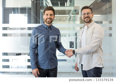 Two happy male business partners closing a successful deal in a corporate office setting, shaking hands and smiling at the camera, symbolizing agreement and collaboration Two happy male business partners closing a successful deal in a corporate office setting, shaking hands and smiling at the camera, symbolizing agreement and collaboration 133379139