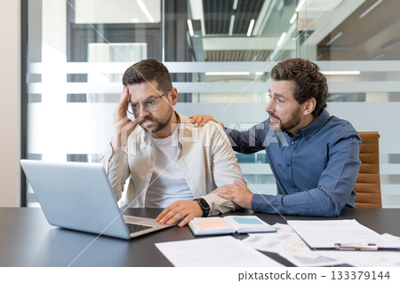 Two male coworkers in a modern office, one offering comfort and support to the other who is stressed, holding his head over a laptop amid paperwork and tension 133379144