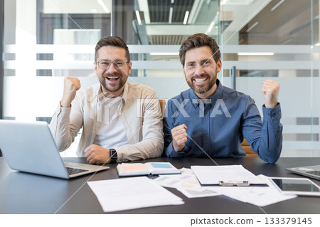 Two cheerful businessmen sitting at a desk with laptops and documents, raising their fists in celebration, happy about achieving a goal or closing a successful deal 133379145