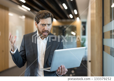 Disappointed business man experiencing stress and confusion while working on a laptop in a modern office hallway, dealing with a difficult technology issue Disappointed business man experiencing stress and confusion while working on a laptop in a modern office hallway, dealing with a difficult technology issue 133379252