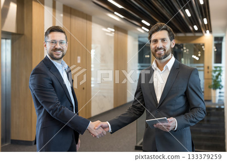 Two smiling businessmen in suits are shaking hands, confirming a successful partnership and agreement in a modern office hallway, one holding a digital tablet Two smiling businessmen in suits are shaking hands, confirming a successful partnership and agreement in a modern office hallway, one holding a digital tablet 133379259