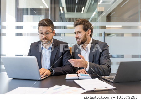Two businessmen discussing project details and analyzing data on a laptop, fostering teamwork and professional collaboration in a modern office environment Two businessmen discussing project details and analyzing data on a laptop, fostering teamwork and professional collaboration in a modern office environment 133379265