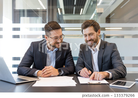 Two smiling adult businessmen sitting at an office desk, reviewing legal papers and signing a new business contract, symbolizing collaboration and agreement Two smiling adult businessmen sitting at an office desk, reviewing legal papers and signing a new business contract, symbolizing collaboration and agreement 133379266