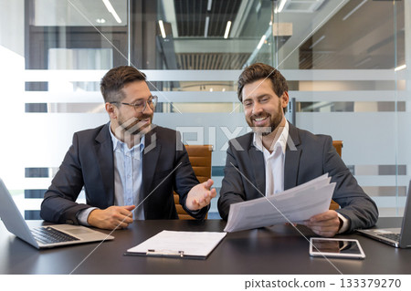 Two smiling businessmen sitting at a modern office table, discussing work documents and collaborating happily during a corporate meeting with laptops and a tablet on the desk 133379270