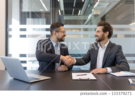 Two smiling businessmen shaking hands, making a partnership agreement after a successful professional meeting in a contemporary office setting, finalizing their collaboration Two smiling businessmen shaking hands, making a partnership agreement after a successful professional meeting in a contemporary office setting, finalizing their collaboration 133379271