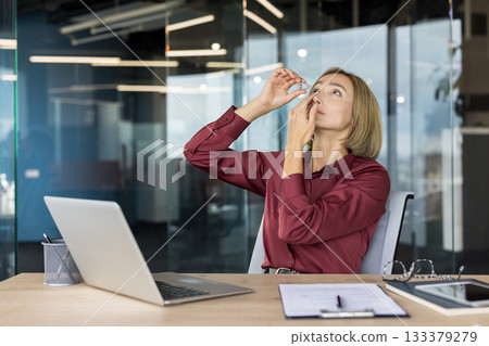 Businesswoman managing eye strain and dry eyes by applying eye drops from a small bottle while working on a laptop at a desk in a contemporary office environment 133379279