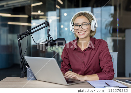 Young woman smiling while recording a podcast or live streaming content, wearing headphones and speaking into a professional microphone at a desk with a laptop and documents Young woman smiling while recording a podcast or live streaming content, wearing headphones and speaking into a professional microphone at a desk with a laptop and documents 133379283