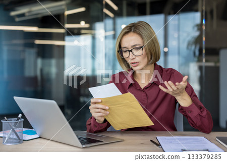 Businesswoman at her desk reading a brown envelope, shocked and frustrated by bad news in a document, showing confusion and stress in a corporate office setting Businesswoman at her desk reading a brown envelope, shocked and frustrated by bad news in a document, showing confusion and stress in a corporate office setting 133379285