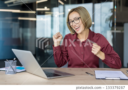 Happy businesswoman in a modern office celebrates online shopping success, holding a credit card and cheering at her laptop after a winning e-commerce transaction and sale 133379287
