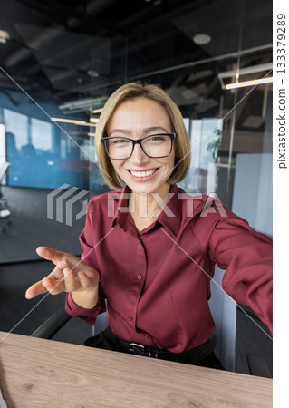 Happy businesswoman wearing eyeglasses and a maroon shirt, smiling and gesturing while participating in a virtual meeting or online conference from her corporate workspace Happy businesswoman wearing eyeglasses and a maroon shirt, smiling and gesturing while participating in a virtual meeting or online conference from her corporate workspace 133379289