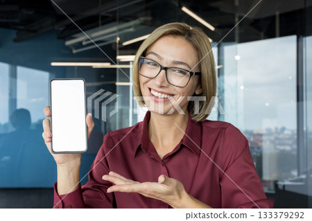 Female professional entrepreneur enthusiastically showing a mobile phone with a white empty screen for advertising or app promotion against a modern office background 133379292