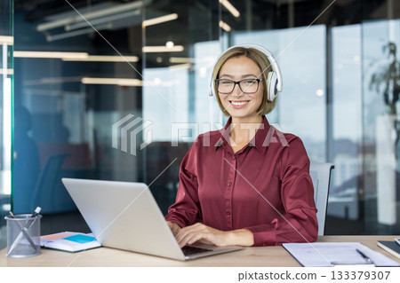 Smiling businesswoman wearing glasses and white headphones, sitting at an office desk and happily working on a laptop, embodying professionalism, concentration, and modern workplace communication Smiling businesswoman wearing glasses and white headphones, sitting at an office desk and happily working on a laptop, embodying professionalism, concentration, and modern workplace communication 133379307