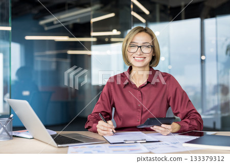 Young female professional accountant working with documents and a calculator at her desk, smiling confidently at the camera, representing success, financial planning, and business management Young female professional accountant working with documents and a calculator at her desk, smiling confidently at the camera, representing success, financial planning, and business management 133379312