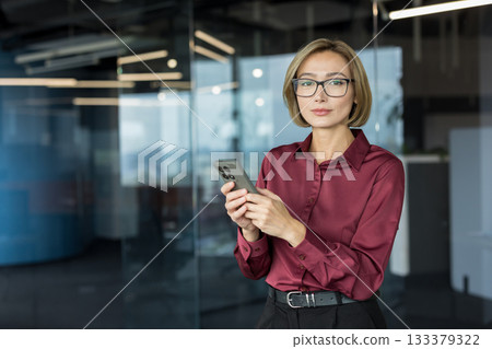 Professional asian businesswoman standing in modern office holding a smartphone, looking at camera while engaging in communication, work, or social networking Professional asian businesswoman standing in modern office holding a smartphone, looking at camera while engaging in communication, work, or social networking 133379322
