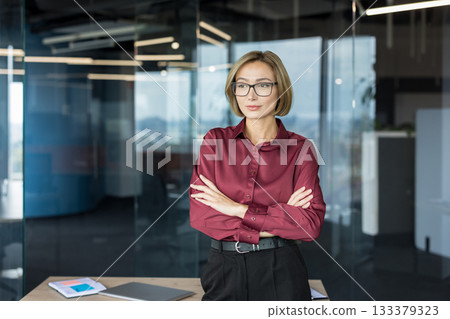 Confident businesswoman with a bob haircut and glasses, wearing a maroon shirt, stands with crossed arms, looking determined inside a contemporary office workplace Confident businesswoman with a bob haircut and glasses, wearing a maroon shirt, stands with crossed arms, looking determined inside a contemporary office workplace 133379323
