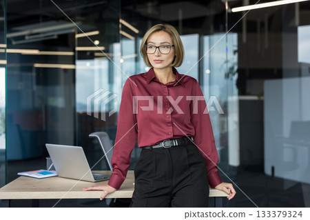 Young professional businesswoman standing confidently by her office desk, contemplating future business strategies within a modern corporate workspace setting Young professional businesswoman standing confidently by her office desk, contemplating future business strategies within a modern corporate workspace setting 133379324