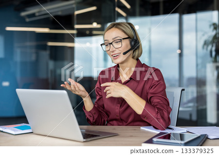 Young businesswoman wearing a headset while actively participating in a video call on her laptop, communicating with clients or team members from a modern office setting Young businesswoman wearing a headset while actively participating in a video call on her laptop, communicating with clients or team members from a modern office setting 133379325