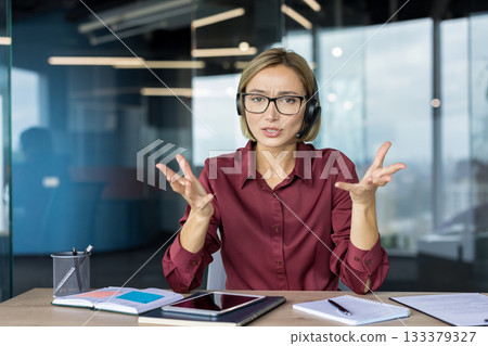 Businesswoman wearing a headset engaging in a video call, expressing confusion and frustration with open hands while working at her office desk in a modern workplace 133379327