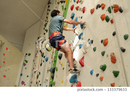 man with safety rope and alpine harness climbing indoor wall with colorful holds 133379633
