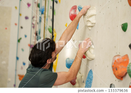 Sporty man climbing with rope and harness on artificial wall in modern fitness center. Sporty man climbing with rope and harness on artificial wall in modern fitness center. 133379636
