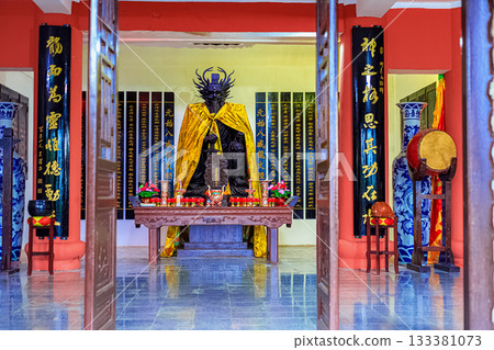 A statue of a dragon-man deity with a yellow veil and sword, surrounded by sacred texts on the walls 133381073