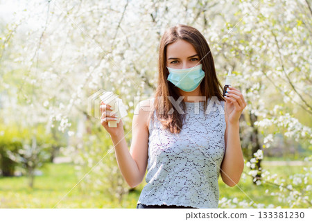 Angry female having allergy standing in blooming spring garden wearing medical mask, holding handkerchief and nasal spray for allergy Angry female having allergy standing in blooming spring garden wearing medical mask, holding handkerchief and nasal spray for allergy 133381230