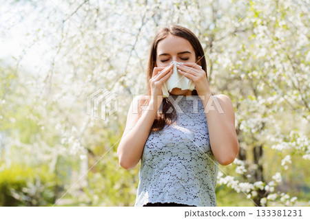 Sneezing young girl with handkerchief among blooming trees in spring park. Concept of allergy Sneezing young girl with handkerchief among blooming trees in spring park. Concept of allergy 133381231