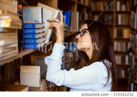 Female college student taking book from shelf in library. Hard-working student looking for a book to find necessary information and prepare report of historical event Female college student taking book from shelf in library. Hard-working student looking for a book to find necessary information and prepare report of historical event 133381232