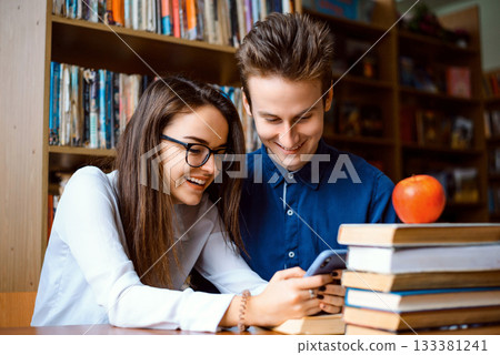 Happy male and female students watching content in their mobile phones, having break of preparing for classes in the library 133381241