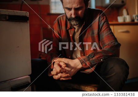 Despaired man sitting by wood stove in aged interior. 133381438