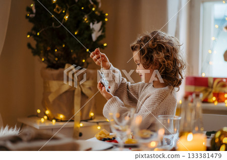 Cheerful small girl indoors at home at Christmas, looking at christmas ornament. Cheerful small girl indoors at home at Christmas, looking at christmas ornament. 133381479