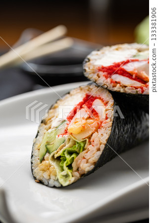Sushi burrito halves with nori, crab, roe and veggies on white plate with soy sauce, close up 133381906